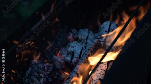 A close-up of burning wood in a barbecue grill. The smoldering embers, crackling wood, and rising flames create a warm, rustic atmosphere.