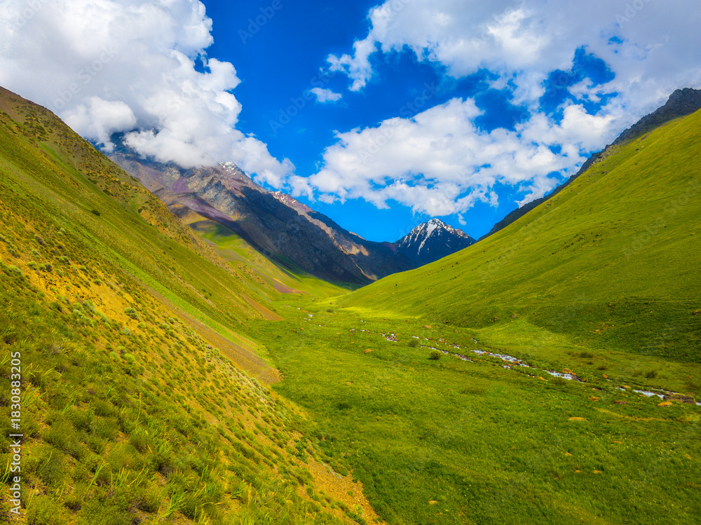 Naklejka premium Majestic mountains rise above a tranquil valley, with a clear stream flowing through the lush greenery. Daylight illuminates the peaceful scene, evoking a sense of serenity and natural beauty.