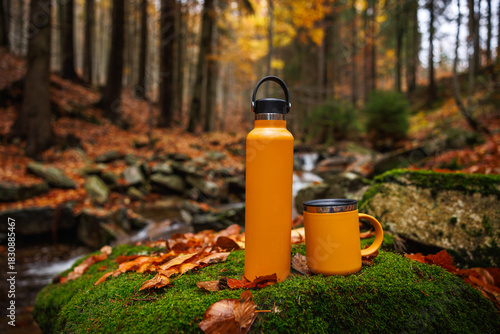 Thermos and travel mug at mountain stream in autumn forest. Hiking equipment. Insulated bottle for keeping hot drink all day