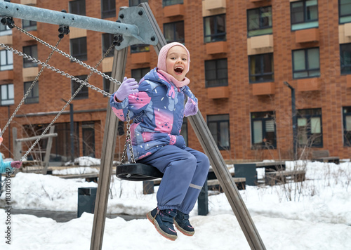Happy girl in snowy playground fun swing and laugh in winter. Children play walking outdoors.