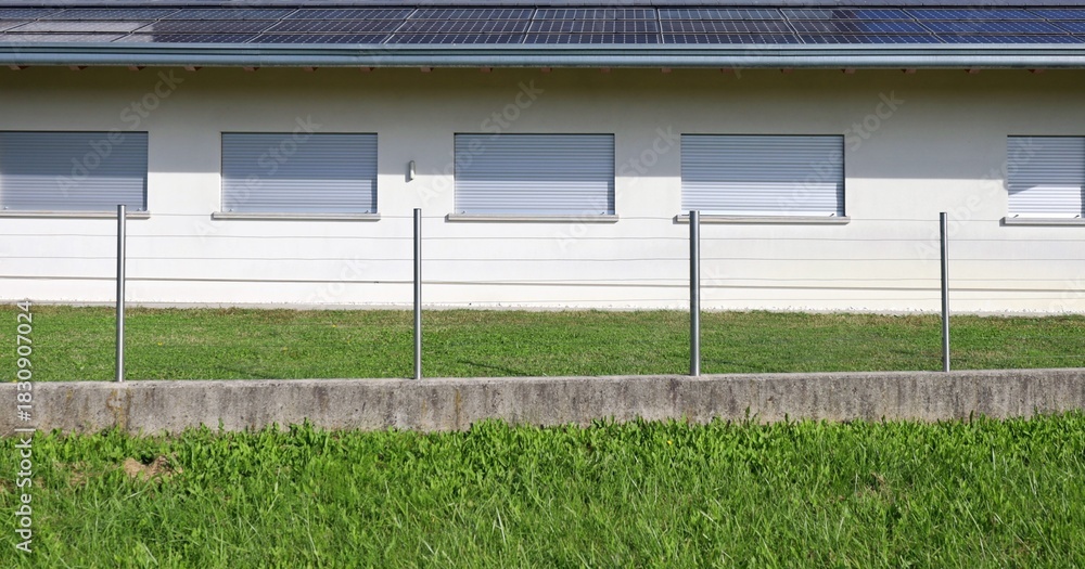 Obraz premium White one-story building with closed shutters and roof panel solar. Meadow and metal post fence in front. Background for copy space.