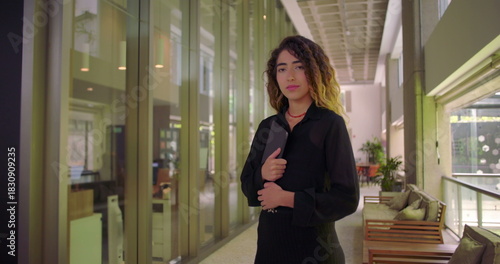 Full body portrait of young woman holding tablet, standing in modern office corridor with confident stance, facing forward in professional workspace