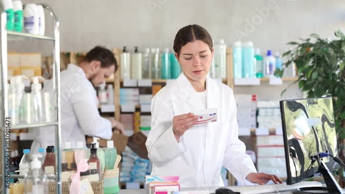 Young woman pharmacist with package of pills, she stands behind the counter near the computer. Pharmacist works in a pharmacy against the background of an male assistant who puts goods on the shelves