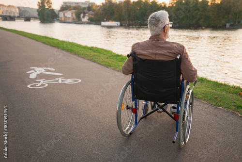 Sad senior man sitting in wheelchair walking on road in city park outdoor, back view. Old paralyzed man in chair for people with disability feel depressed. Lonely elderly sick man sits on wheelchair