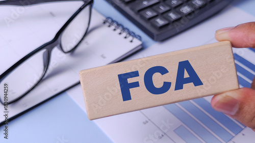 FCA (Financial Conduct Authority) Concept: Wooden Block with Abbreviation Held Over Financial Chart, Calculator, and Notebook