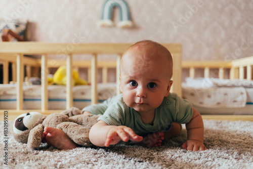 A cute baby lying on their stomach on a bed, looking forward with a curious expression. Warm natural light and a cozy home environment create a tender and intimate family scene.