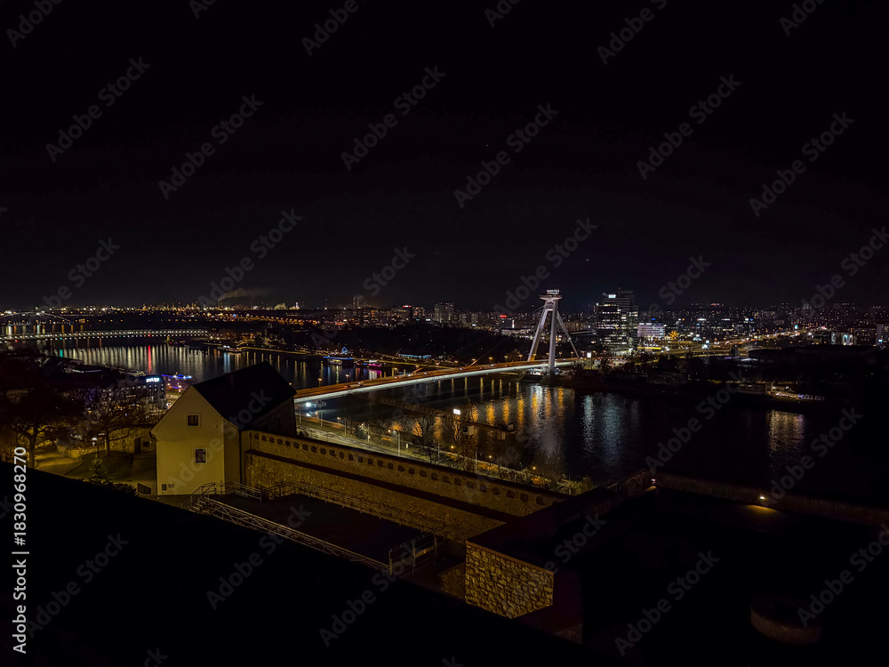 Fototapeta premium Illuminated SNP Bridge with its UFO tower rises over the Danube River at night, showcasing Bratislava’s modern cityscape and waterfront lights.