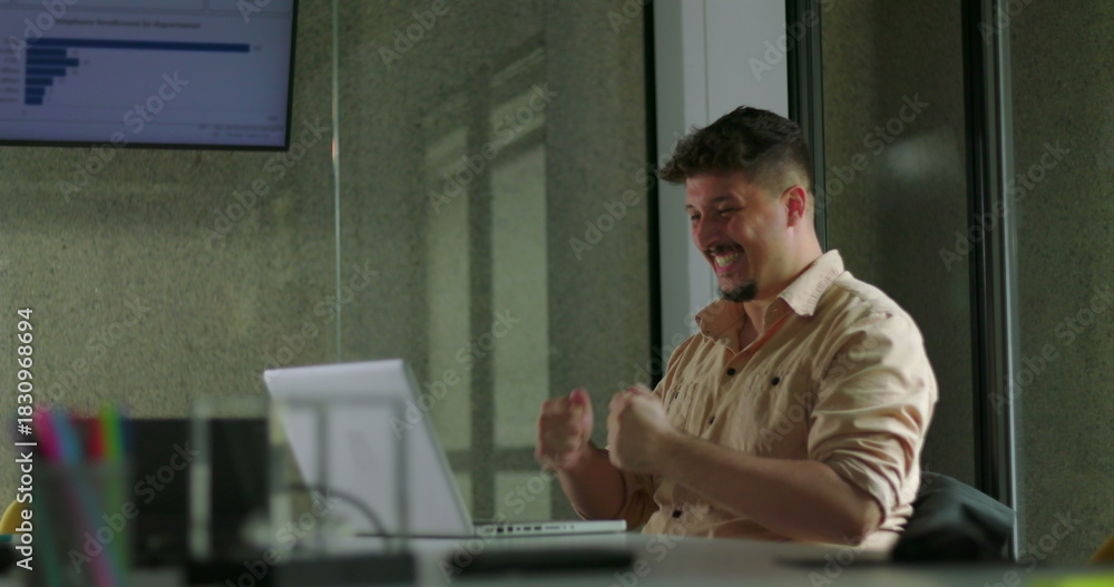 Fototapeta premium Happy young man celebrates success alone at laptop with double fist pump during solo office work session