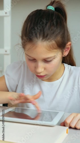 Young girl in white shirt focused on playing a game on tablet using her fingers while sitting at desk in bright indoor environment