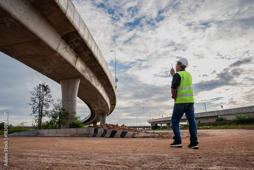 A male civil engineer or site supervisor, wearing a white hard hat, and a neon vest, stands alone on a vast dirt field. The massive concrete structure of a highway overpass dominating the background.