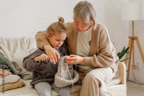 In a cozy living room, a grandmother is guiding her young granddaughter as they knit a hat together. Their shared moment feels warm and joyful