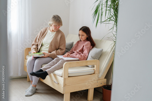A grandmother and her granddaughter sit together on a comfortable sofa, enjoying reading books in a bright and inviting living room