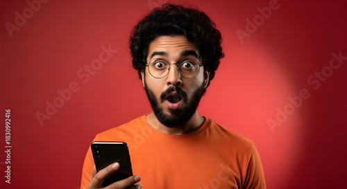 Surprised Man Using Smartphone: Astonished Expression of a Young Adult Reacting to Breaking News or Unexpected Online Content on Mobile with a Red Backdrop