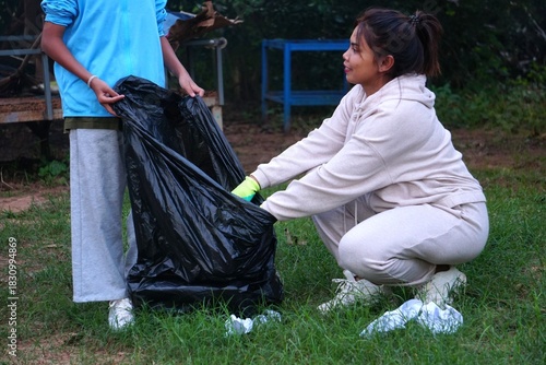 The woman is squatting, using gloves to collect trash and bending down to place it into the large black trash bag held by the girl.