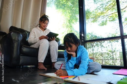 Mother sitting on a couch using a tablet, with her daughter sitting on the floor concentrating on her drawing.