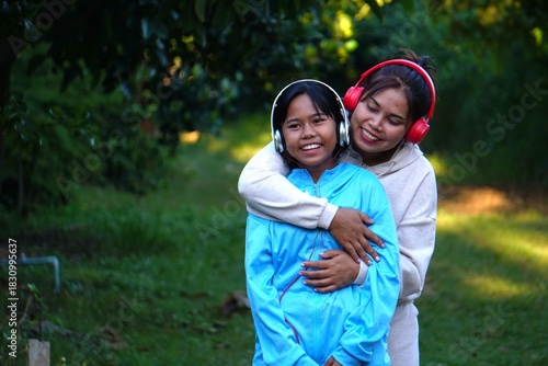 The mother hugs her daughter from behind, and both are smiling happily and wearing headphones.