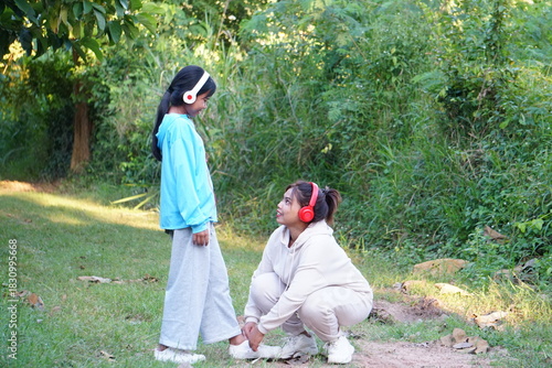 The mother is crouching down to tie her daughter's shoelaces in the park.