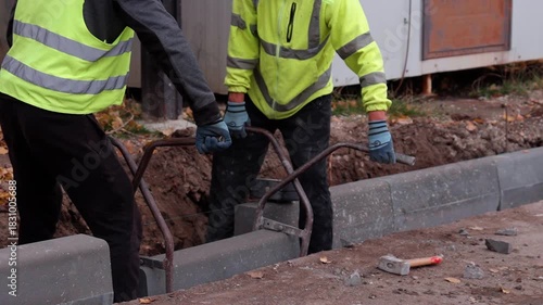 Construction workers in safety gear are installing concrete curbs along a roadway, showcasing teamwork and precision, with a focus on the lifting action and equipment used