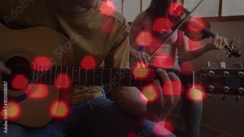 Camera pulling back, guitarist strumming and violinist bowing, rehearsing music under red bokeh