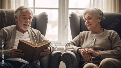 Senior man reading a book aloud to his wife in comfortable armchairs. Elderly couple sharing a moment of companionship at home
