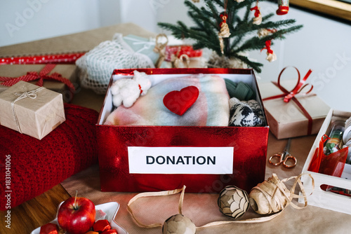 Red box labeled DONATION filled with soft items and heart sits on table surrounded by gifts and small Christmas tree. Holiday donations, Christmas charity, festive giving, spreading kindness.