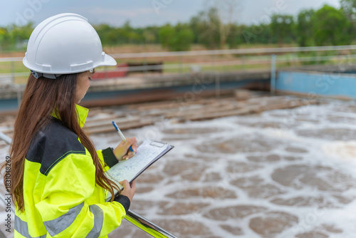 Professional Asian female environmental engineer conducting an inspection and recording operational data on a clipboard at a large industrial water treatment plant sewage facility outdoors.