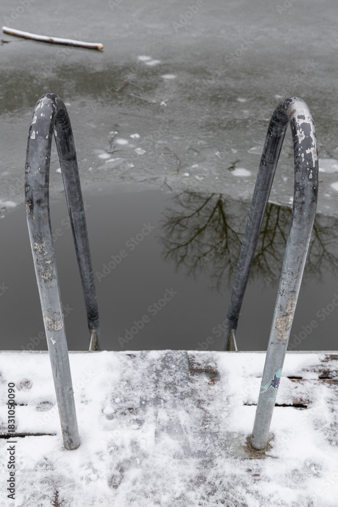 Fototapeta premium Metal ladder handrails entering a partially frozen water pond with snow covered deck. Horejsi rybnik in prague 9