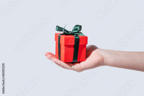 A hand holding a small red gift box with an emerald ribbon on an isolated light background