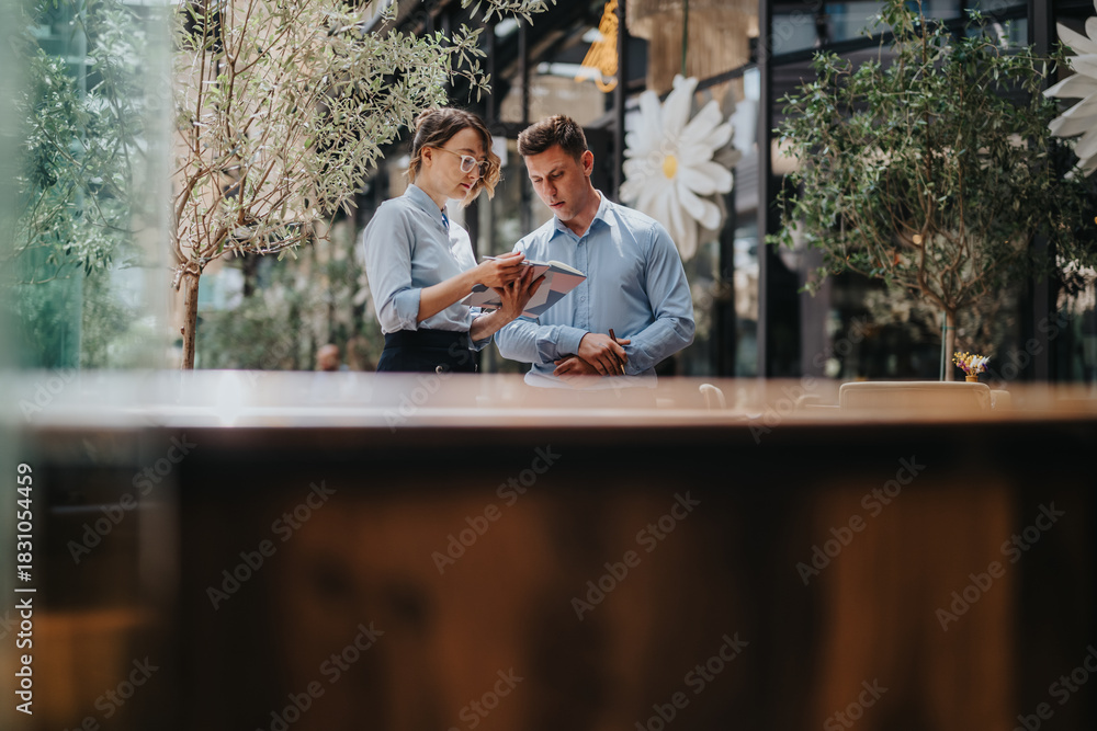 Obraz premium Two colleagues in light blue shirts discuss ideas while looking at a tablet in a stylish indoor space. Greenery and decor create a calm, collaborative, modern atmosphere.