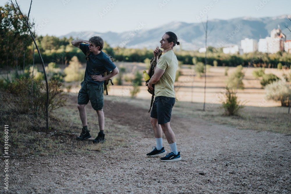 Fototapeta premium Two men with backpacks stand on a gravel path in an open park with mountains in the distance. One looks ahead while the other shields his eyes, sharing a quiet moment before hiking.