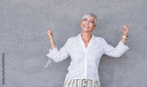 Excited beautiful adult woman with closed eyes and clenched fists, against gray background - wall. Yes concept. Good news