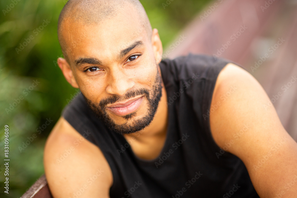 Fototapeta premium Confident young man with shaved head and beard looking at the camera while relaxing on a bench