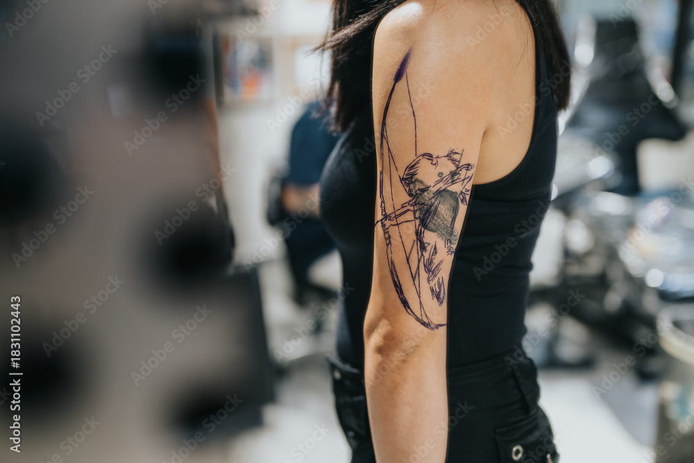 Fototapeta premium A woman in a black tank top shows a bold archer bear tattoo on her upper arm. The sketchy design features dark lines and a dynamic bow pose, suggestive of inked art in progress.