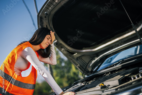 A young woman wearing a safety vest stands in front of her car with the hood up, holding her head, facing a car breakdown situation.