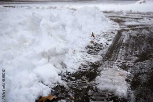 A snow pile lines the edge of a concrete driveway or lot surface marked with tire tracks. The ground shows signs of melting and refreezing, with slushy and icy patches. Winterdienst backdrop. 