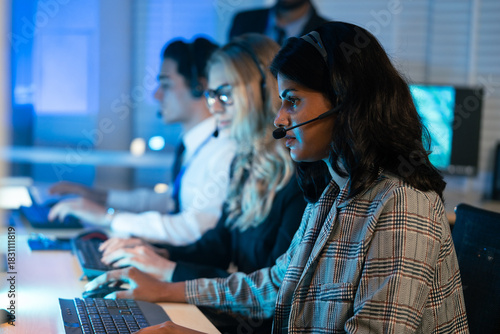 Customer service representative using a headset, working late in a modern, dark office with computer monitors displaying data and analytics. Call center operations, B2B support, technical assistance