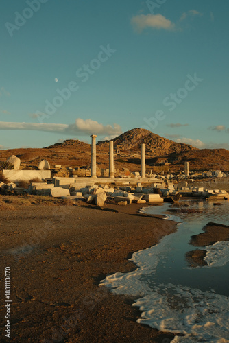 Coastal view of Delos island featuring ancient Greek ruins, standing columns, scattered marble blocks and calm waves illuminated by warm sunset light.