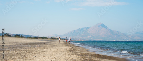 Fototapeta Naklejka Na Ścianę i Meble -  A wild seaside beach with a group of tourists strolling and jogging along the shoreline. Mountains in the background. Tyrrhenian Sea. Sicily.
