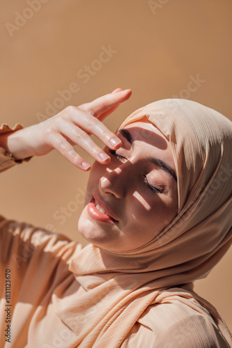 Schilderij op canvas Young woman wearing a light-colored hijab gently touches her face while basking