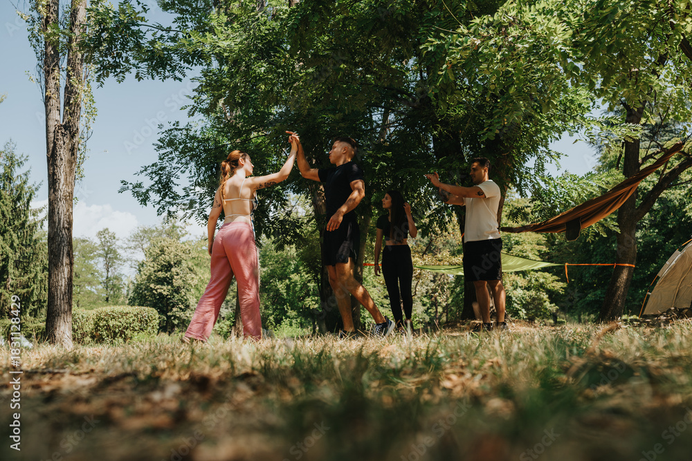 Fototapeta premium A joyful group of friends and family share a high-five moment at a sunny campsite. Kids and adults relax under green trees with tents and a hammock nearby, conveying outdoor leisure and togetherness.