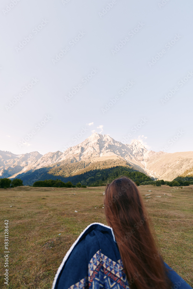 Naklejka premium Mountain landscape with woman in meadow, outdoor hiking scene showing long hair and jacket from back, scenic peak and wide sky, travel and adventure mood in natural wilderness.