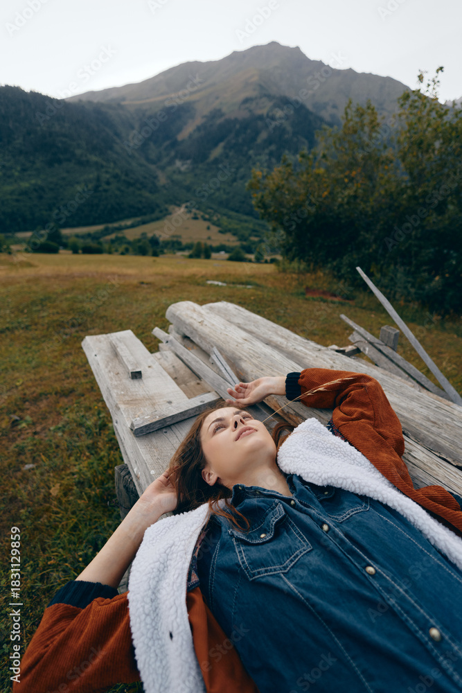 Fototapeta premium woman outdoors in meadow and mountains lying on wood planks, portrait of relaxation under open sky, casual denim jacket and cozy coat in peaceful natural landscape