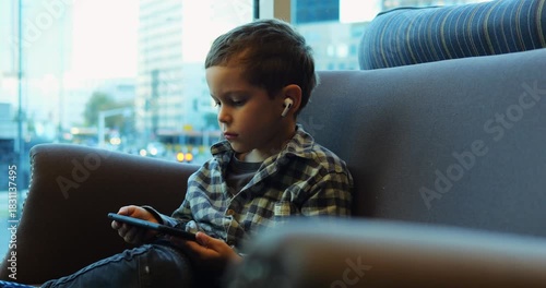 Young preschooler in earbuds deeply engrossed in phone while comfortably seated on sofa. Immersive experience with digital content in relaxed indoor setting