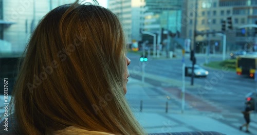 Woman gazes contemplatively in large window observing cityscape with buildings and traffic. Posture suggests moment of observation of bustling city environment