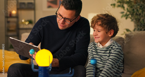 Son is spending time with dad men are sitting on couch playing with a model of planetary system guy is explaining to boy about construction of cosmos using toy as example he is naming planets.