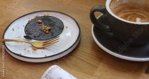 Plate with cake crumbles and half of coffee cup with bill on cafe wooden table. Leftovers of lunch with dessert and cappuccino in coffeeshop