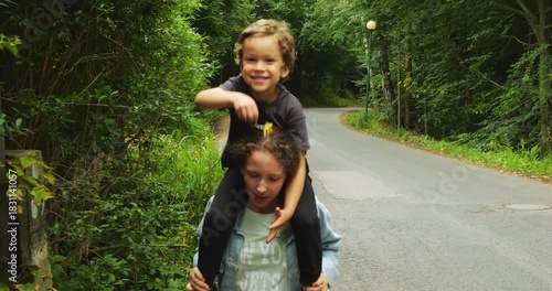 Child smiles brightly while sitting on sister shoulders walking on path surrounded by green trees. Scene conveys joyful family moment during outdoor stroll