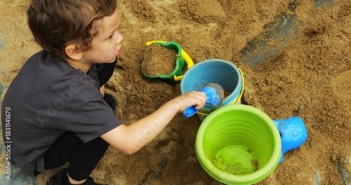 Preschooler plays in sandbox surrounded by sand toys including buckets and shovels. Child actively digs in and, with dinosaur skeleton on surface