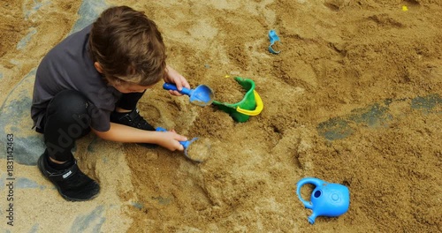 Boy deeply engaged in playing in sandbox utilizing shovels to unearth areas. Dinosaur skeleton beneath sand indicates archaeological excavation theme
