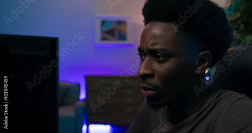 Close up shot of young man's face, professional gamer, sits comfortably in chair, bent over computer, playing video games, focused on devising strategy, room lit with purple, blue led lights.